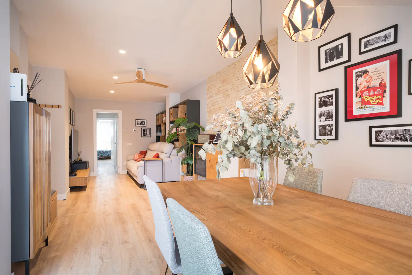 A bright, modern living and dining area with a wooden table, chairs, and geometric pendant lights. Gallery wall with framed art.