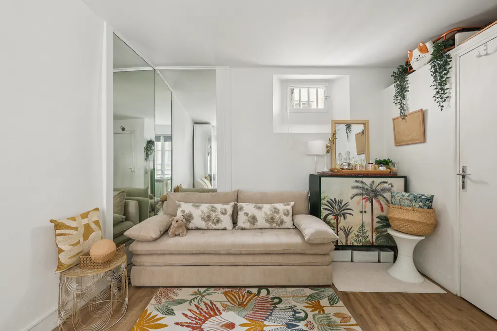 Cozy living room with beige sofa, floral pillows, and colorful rug. Mirrored wall reflects the space. A cabinet with palm tree art adds charm.