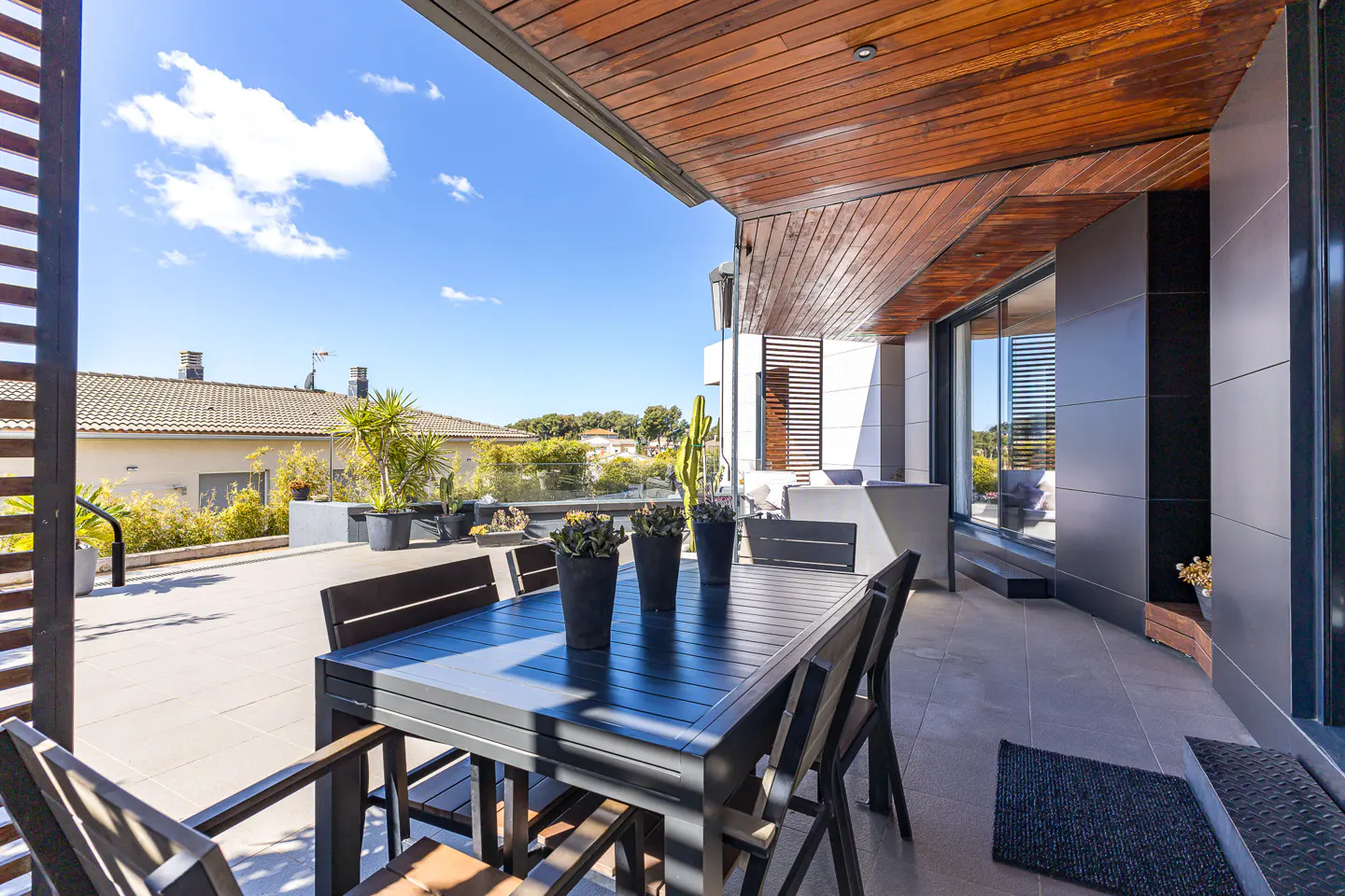 Outdoor patio with a dark table and chairs under a wood ceiling. Potted plants and a view of houses in the background.