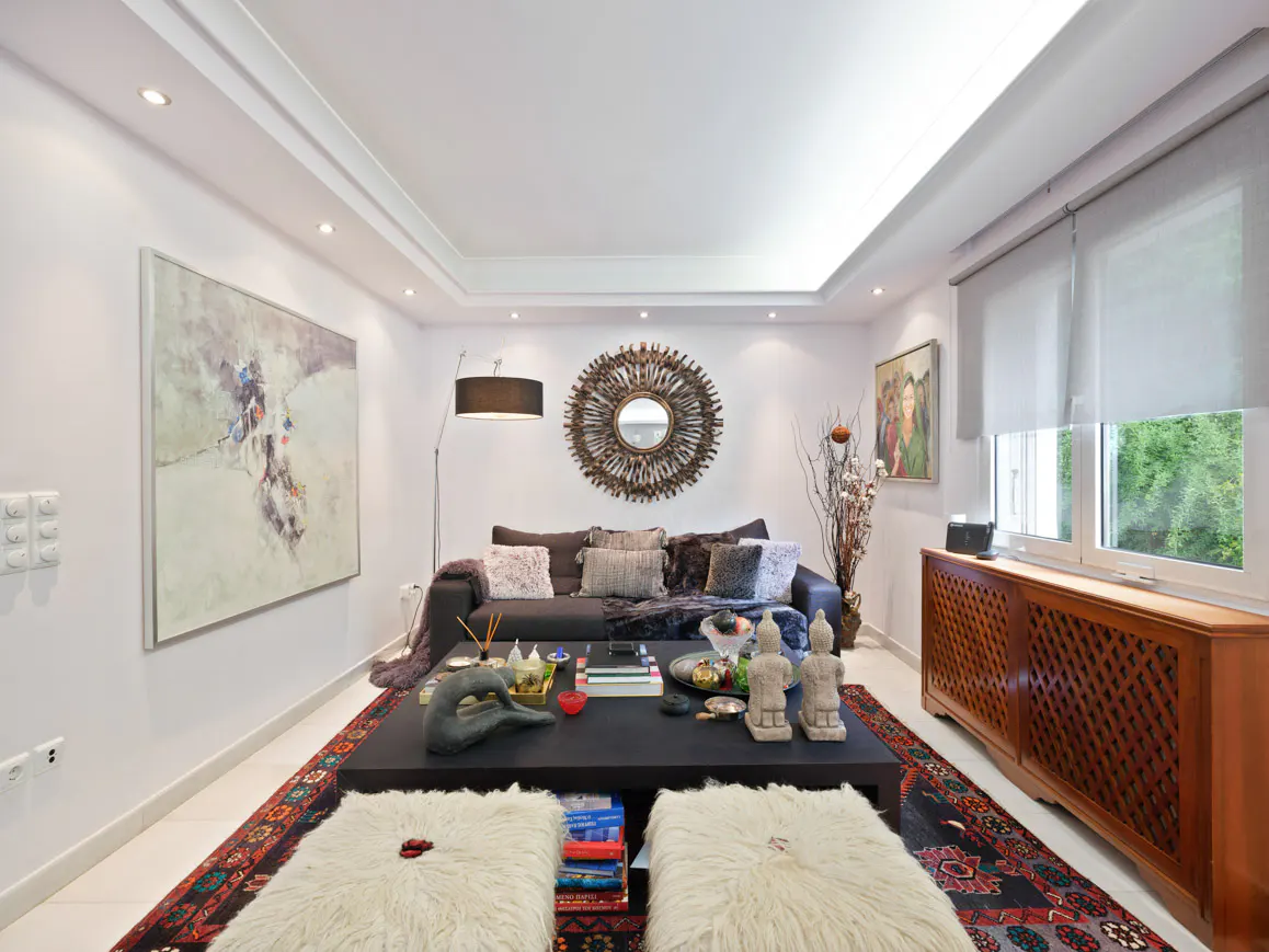 Modern living room with a dark sofa, black coffee table, two white fur ottomans, and a decorative sunburst wall mirror.