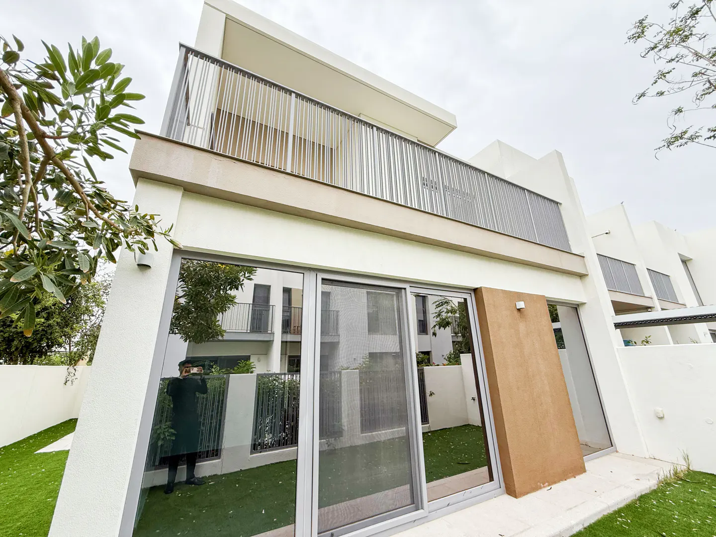 Modern two-story white house with a balcony and large sliding glass doors. Green lawn and trees surround the building.
