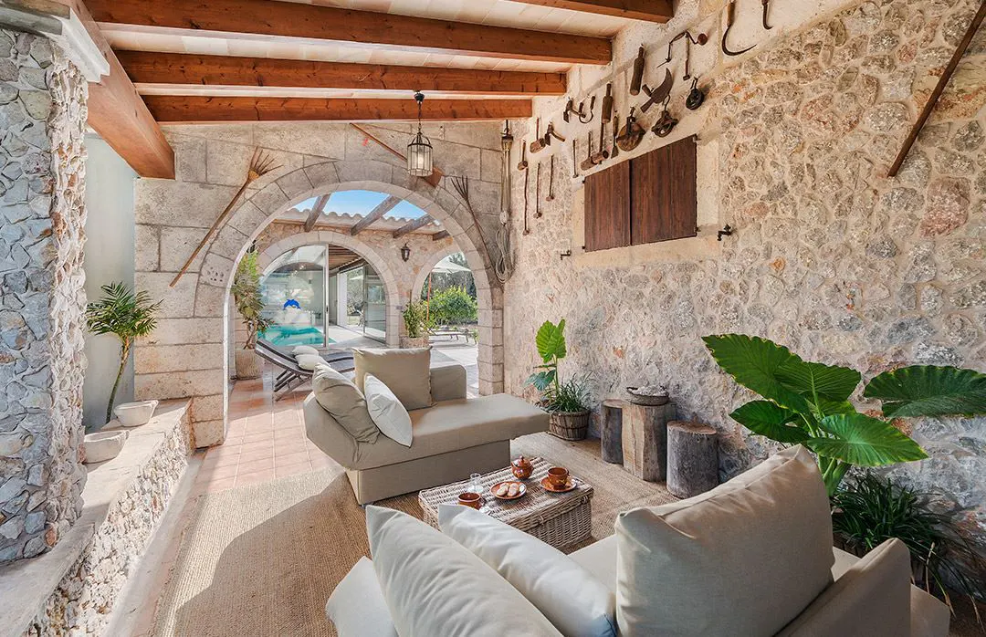 Stone patio with arched openings, beige sofas, and rustic decor. Farm tools hang on the stone wall. A pool is visible through the arches.