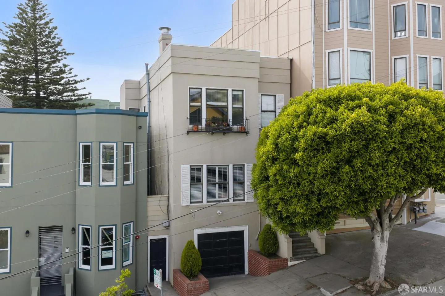 A three-story, light-gray house with a black garage door and a green tree on the right.