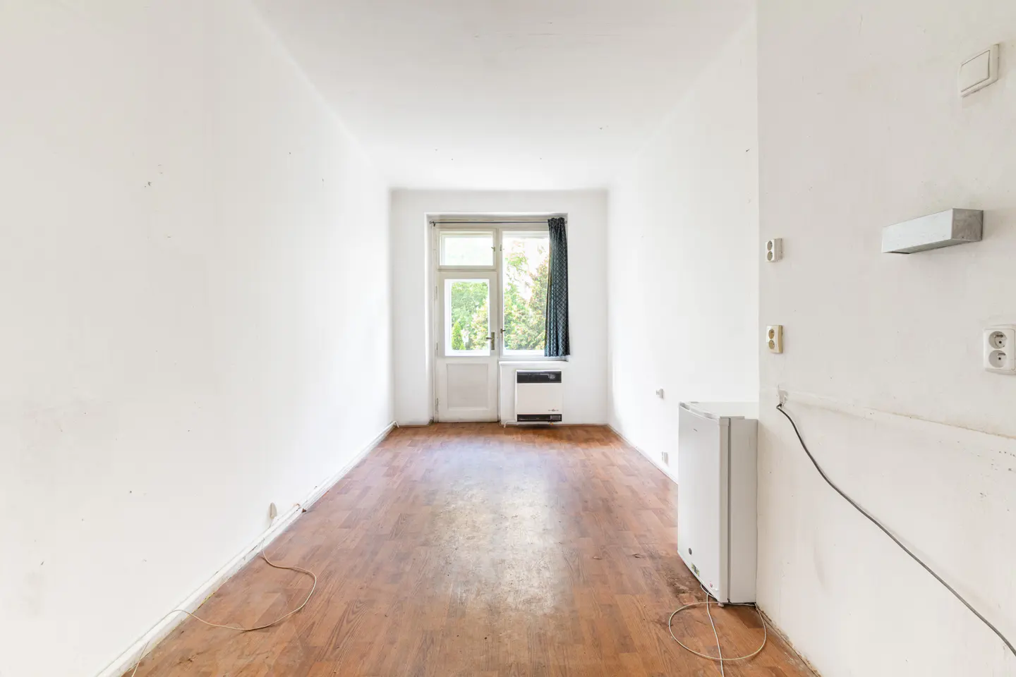 Empty room with white walls, wood-look flooring, a small white refrigerator, and a glass door leading to the outdoors.