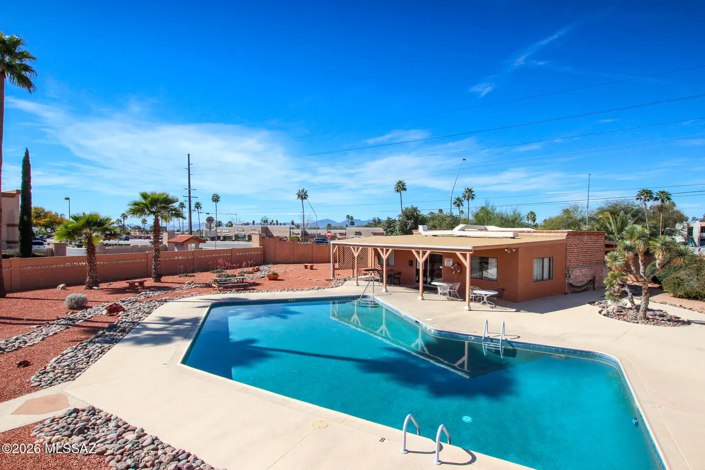 Outdoor pool with clear blue water, surrounded by a beige concrete deck and red gravel. Palm trees and a building with a covered patio are visible.