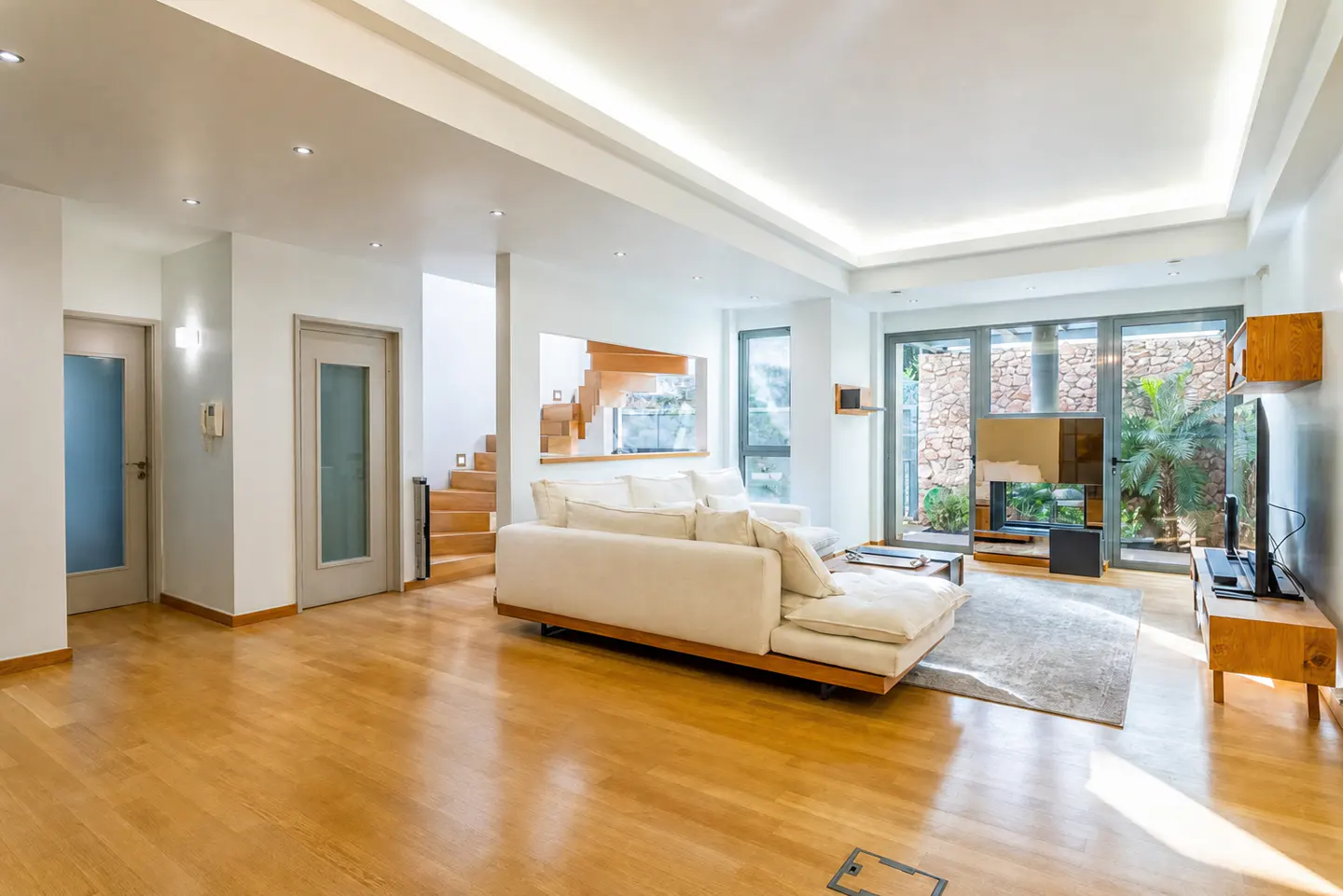 Modern living room with white sofa, hardwood floors, recessed lighting, and large windows overlooking a stone patio.