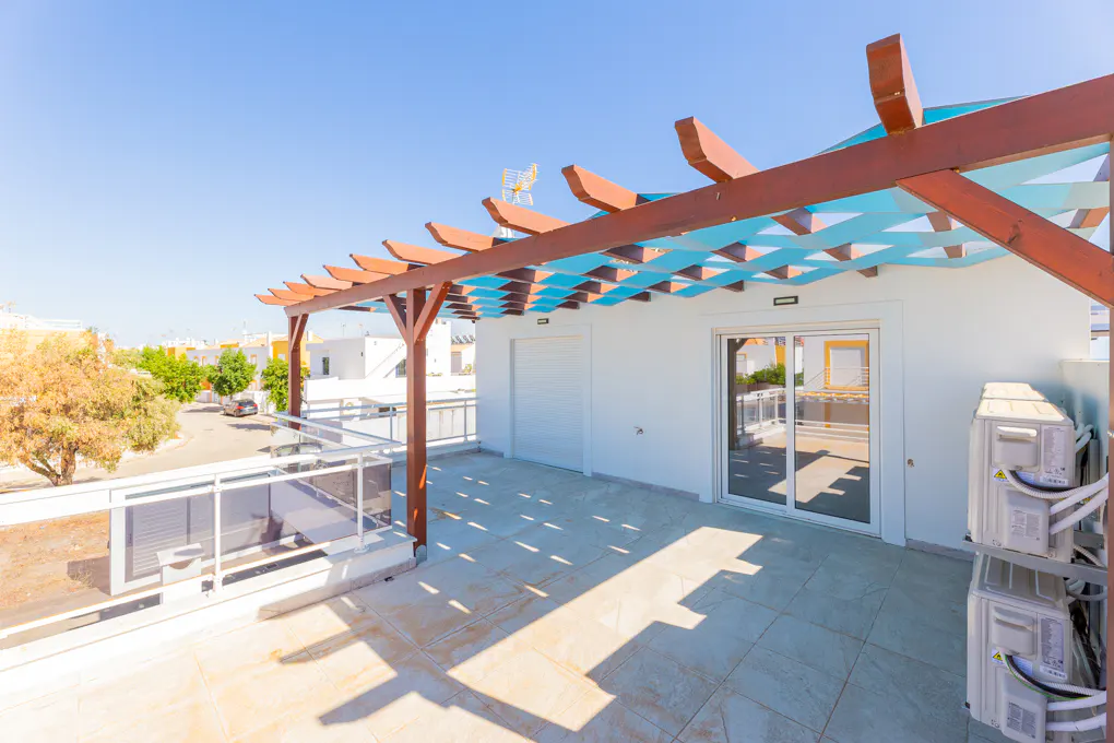 A rooftop patio with a brown pergola and blue lattice, white walls, and a glass railing overlooking a street.