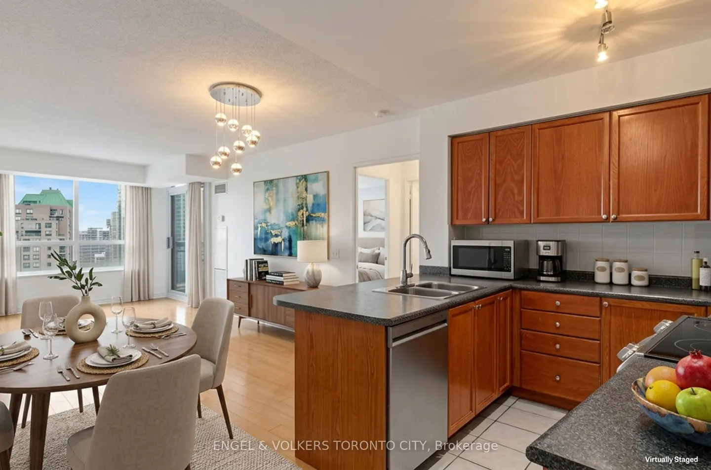 Bright condo interior with wood cabinets, stainless steel appliances, and a dining table set for a meal. City view visible through large windows.