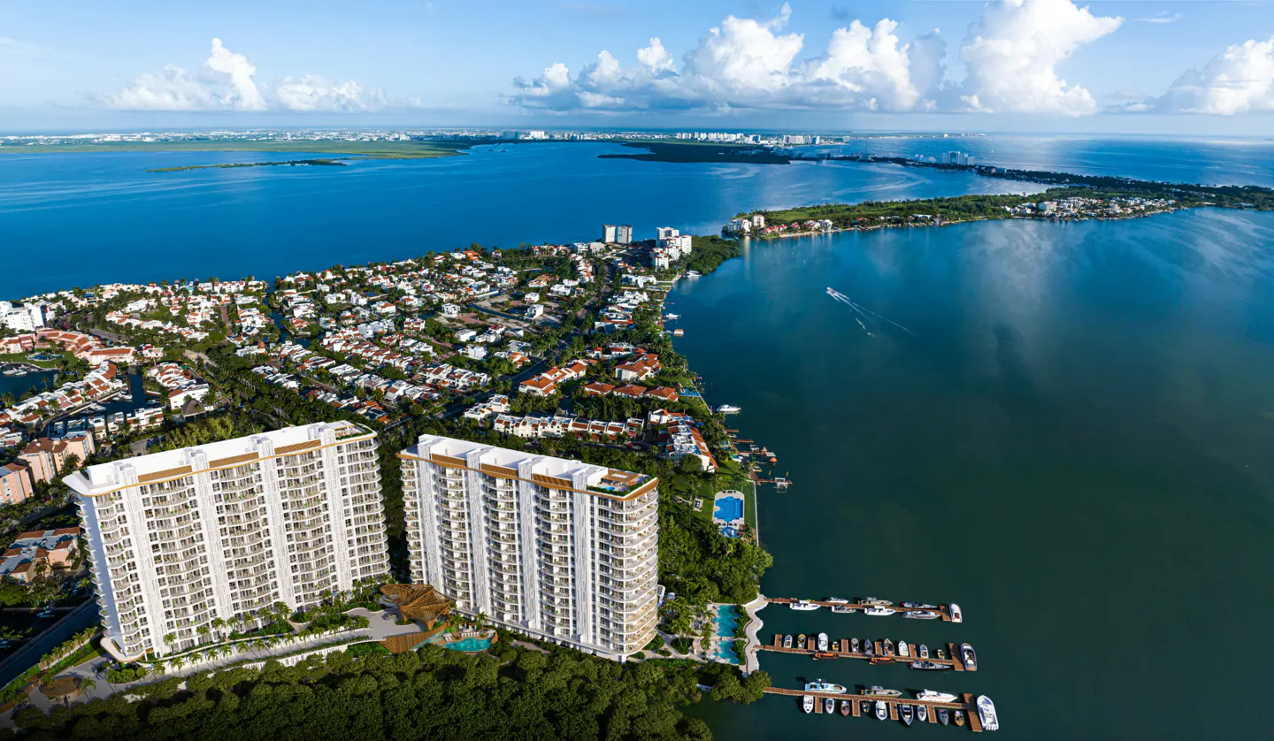 Aerial view of two white condo buildings with a marina on the water, surrounded by lush greenery and a blue bay under a partly cloudy sky.
