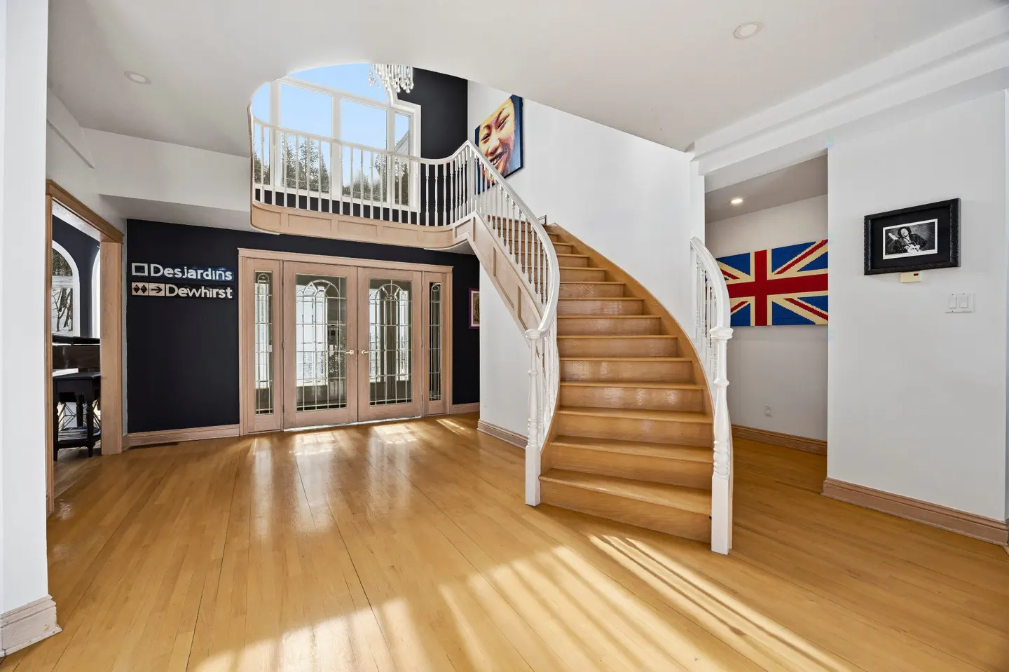Bright foyer with wood floors, a curved staircase with white railings, and double doors. Artwork and a British flag add character.