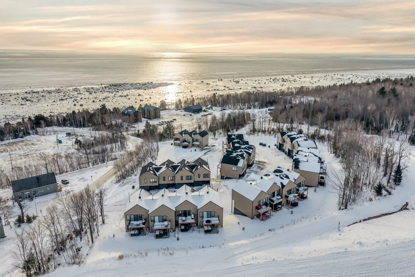 Aerial view of tan townhouses with snow-covered roofs, surrounded by snow-covered ground and bare trees, near a body of water.
