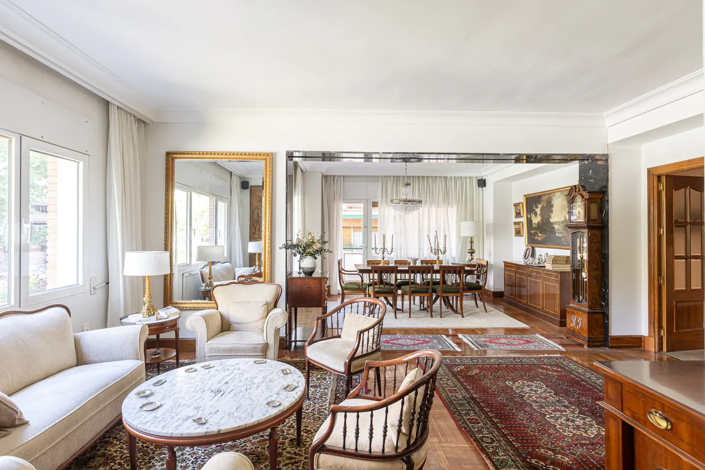 Elegant living room with a marble coffee table, cream armchairs, a dining area, and a large ornate gold-framed mirror.