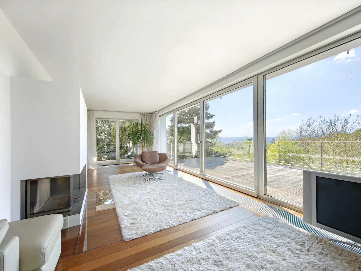 Bright living room with hardwood floors, white walls, and large sliding glass doors to a patio with a view. A brown leather chair sits on a white shag rug.