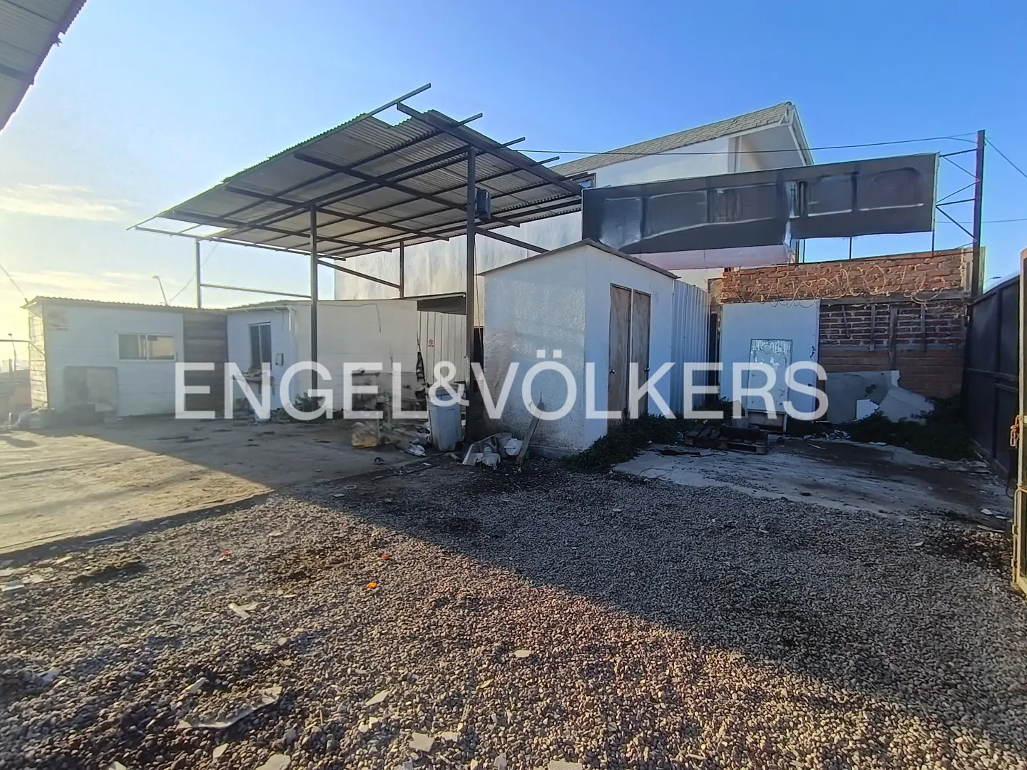 Exterior view of a property with a gravel yard, sheds, and a metal awning. The sky is blue.