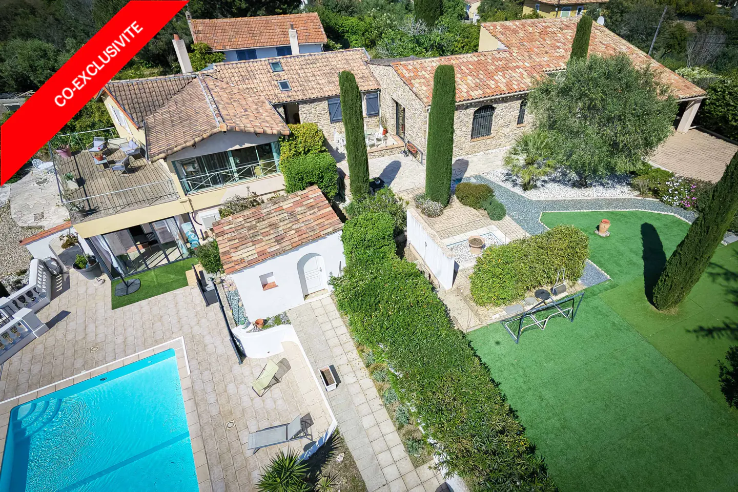 Aerial view of a Mediterranean-style home with a red tile roof, pool, green lawn, and lush landscaping. "Co-Exclusive" banner in the corner.