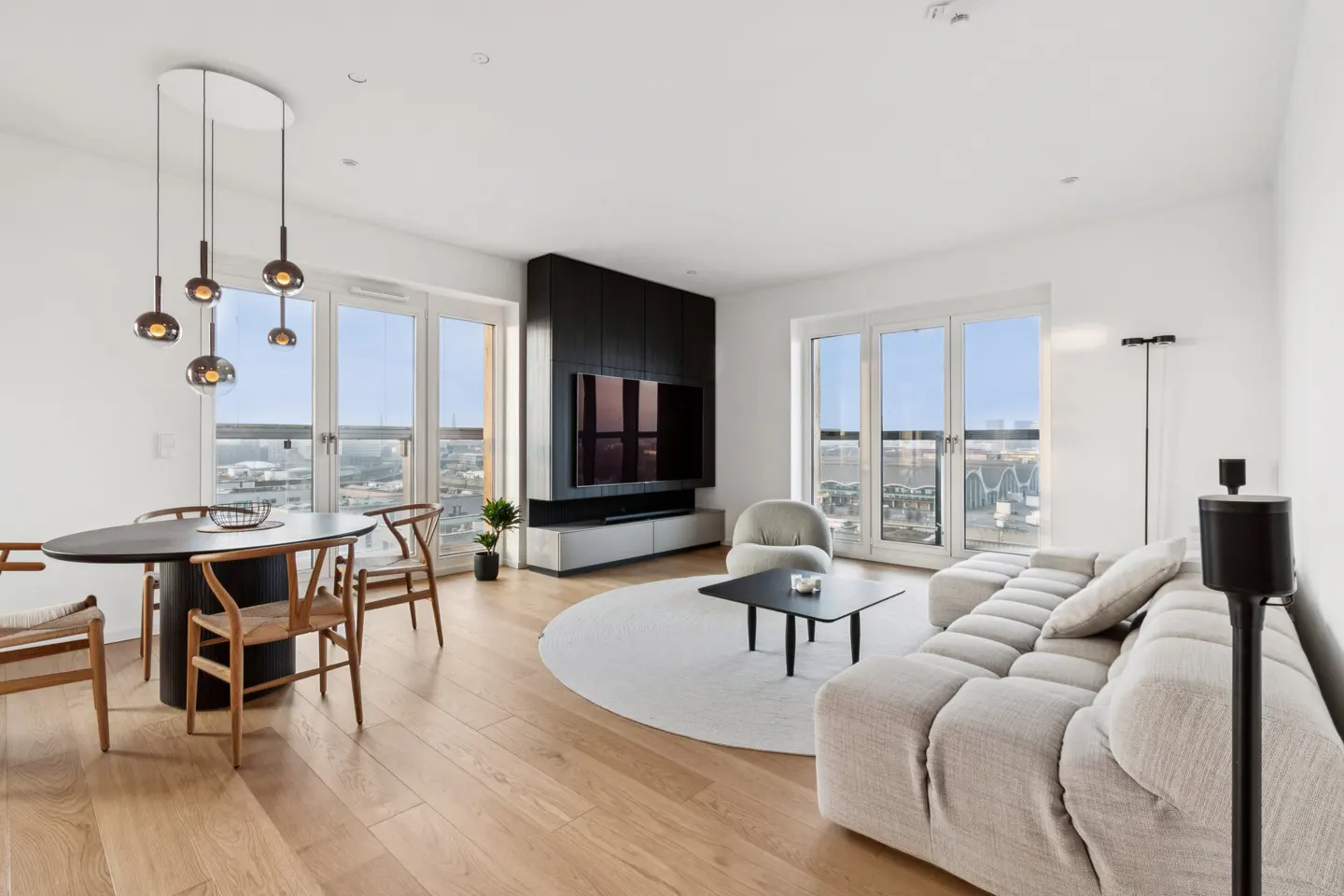 Bright, modern living room with wood floors, white walls, and large windows overlooking a city. A gray sofa and black dining set furnish the space.