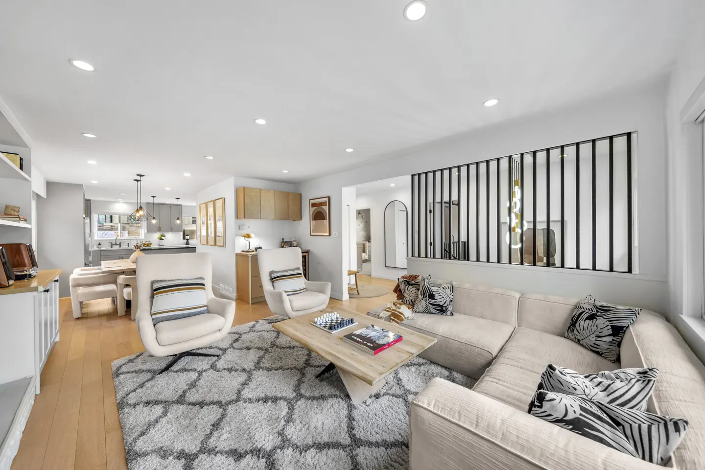 Bright, open-concept living room with white walls, light wood floors, and a gray patterned rug. Two cream armchairs face a beige sectional sofa. A kitchen is visible in the background.