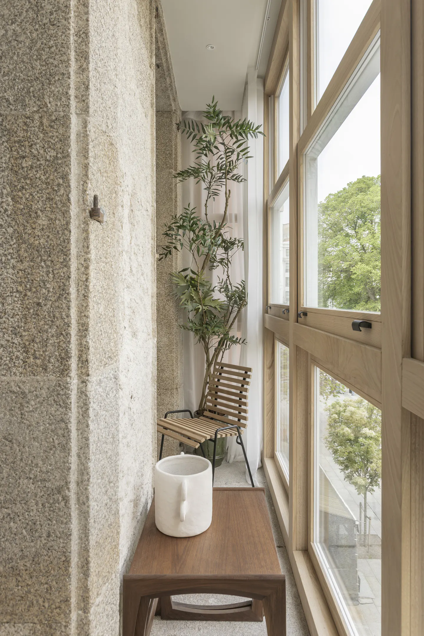 A modern balcony with a stone wall, wooden table, chair, and large windows overlooking greenery. A small tree sits behind the chair.