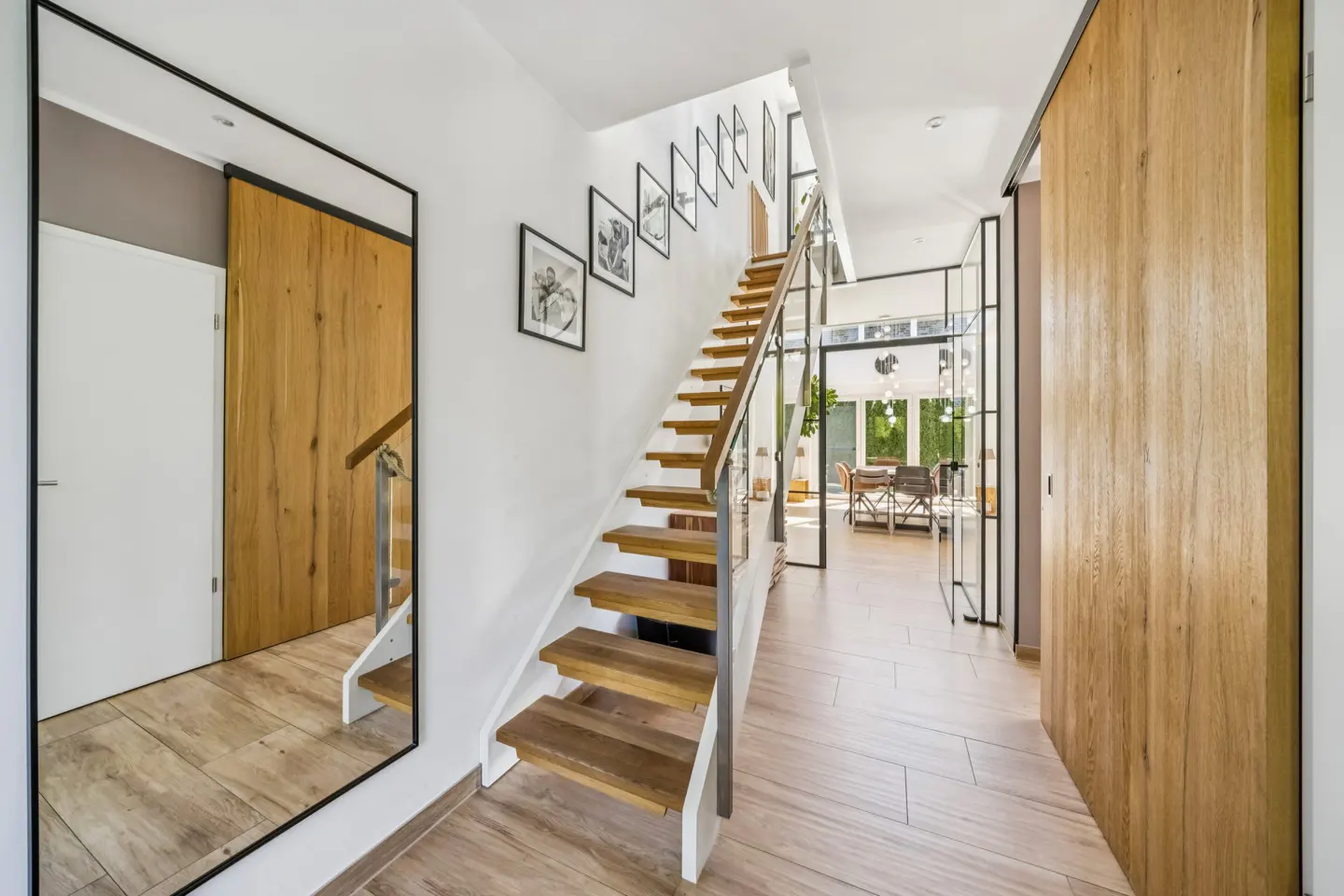 Bright hallway with wood floors, stairs, and a large mirror. Black framed pictures hang on the white wall. A dining room is visible in the background.