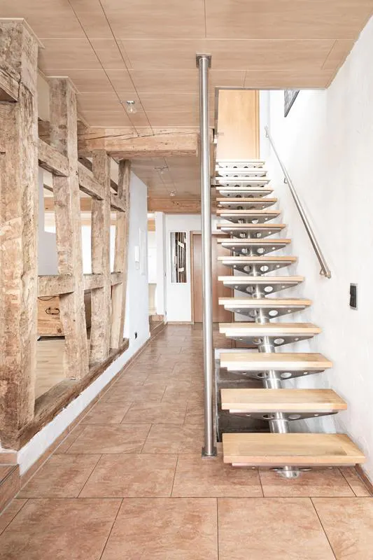 Interior view of a home with a modern staircase, wood beams, and tile flooring. The staircase has metal supports and a handrail.