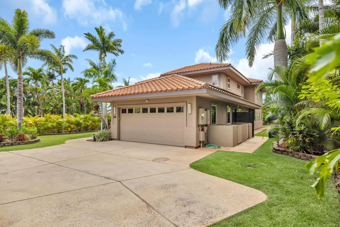 Tan two-story house with a tile roof, large garage, and concrete driveway, surrounded by lush palm trees and green lawn.