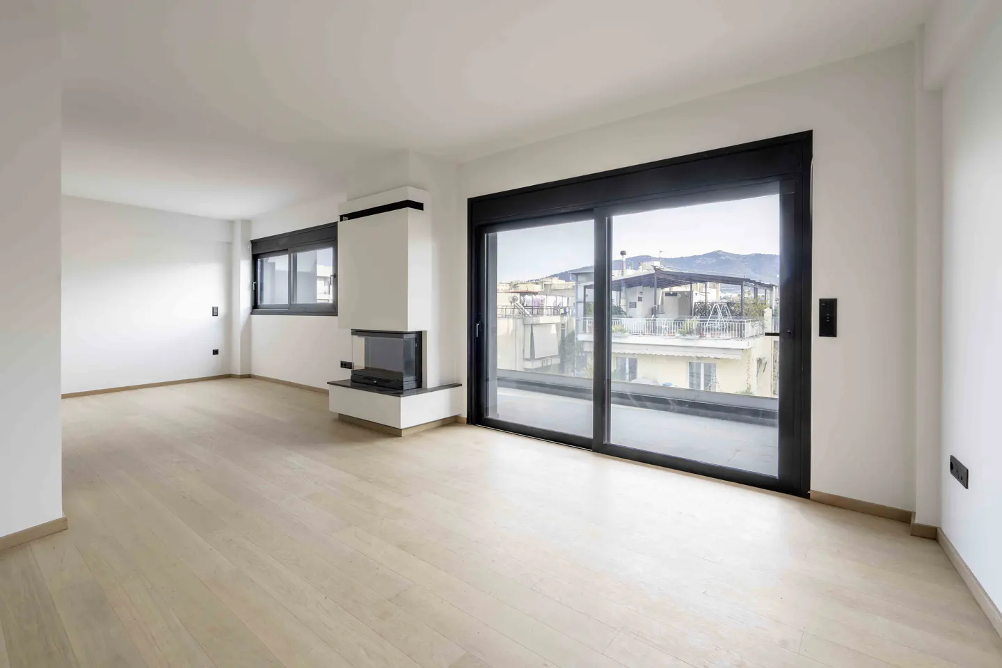 Bright, empty living room with light wood floors, white walls, and a modern white fireplace. A large sliding glass door offers a city view.
