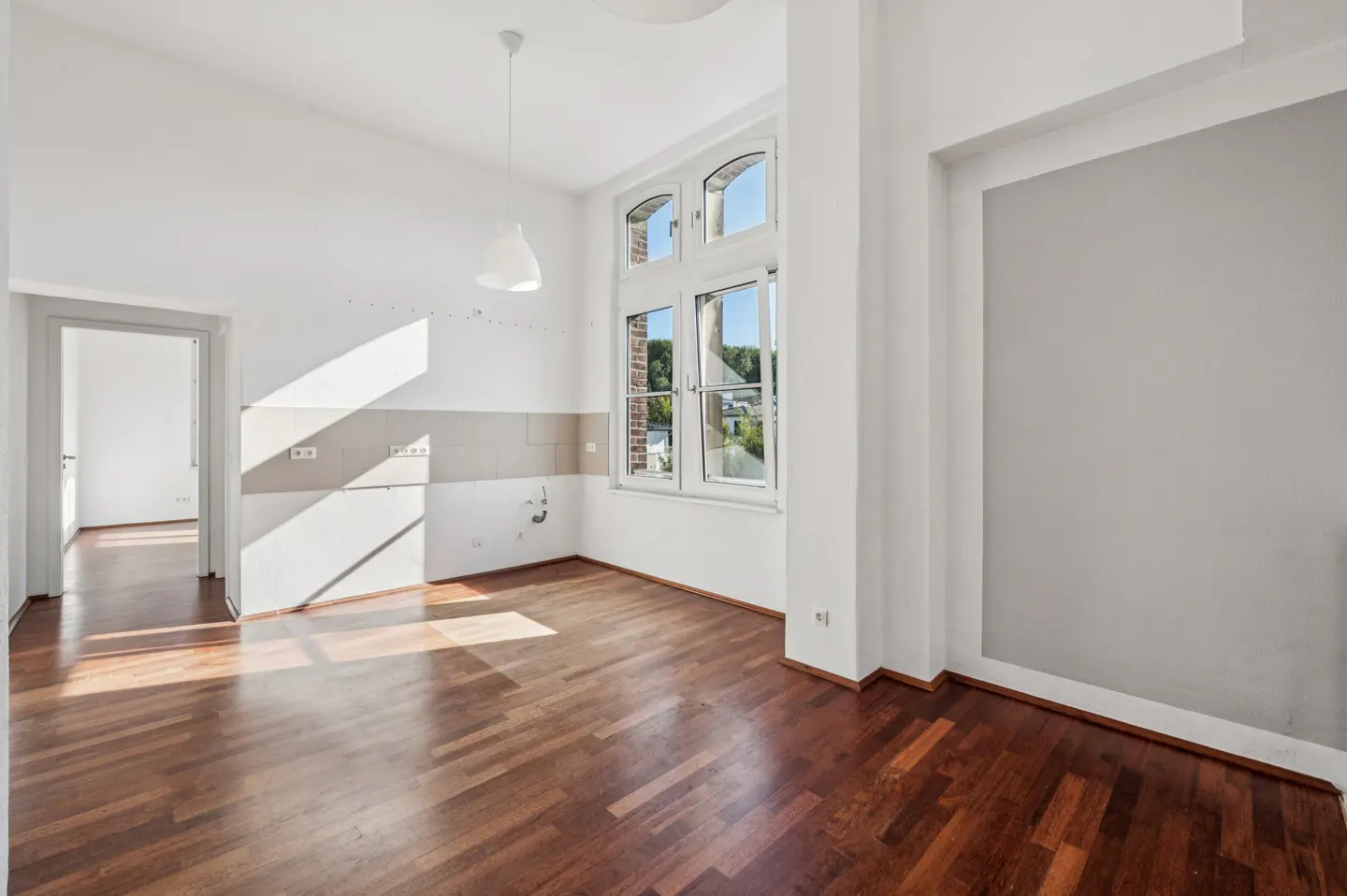 Bright, empty room with hardwood floors, white walls, and a large window. A doorway leads to another room.