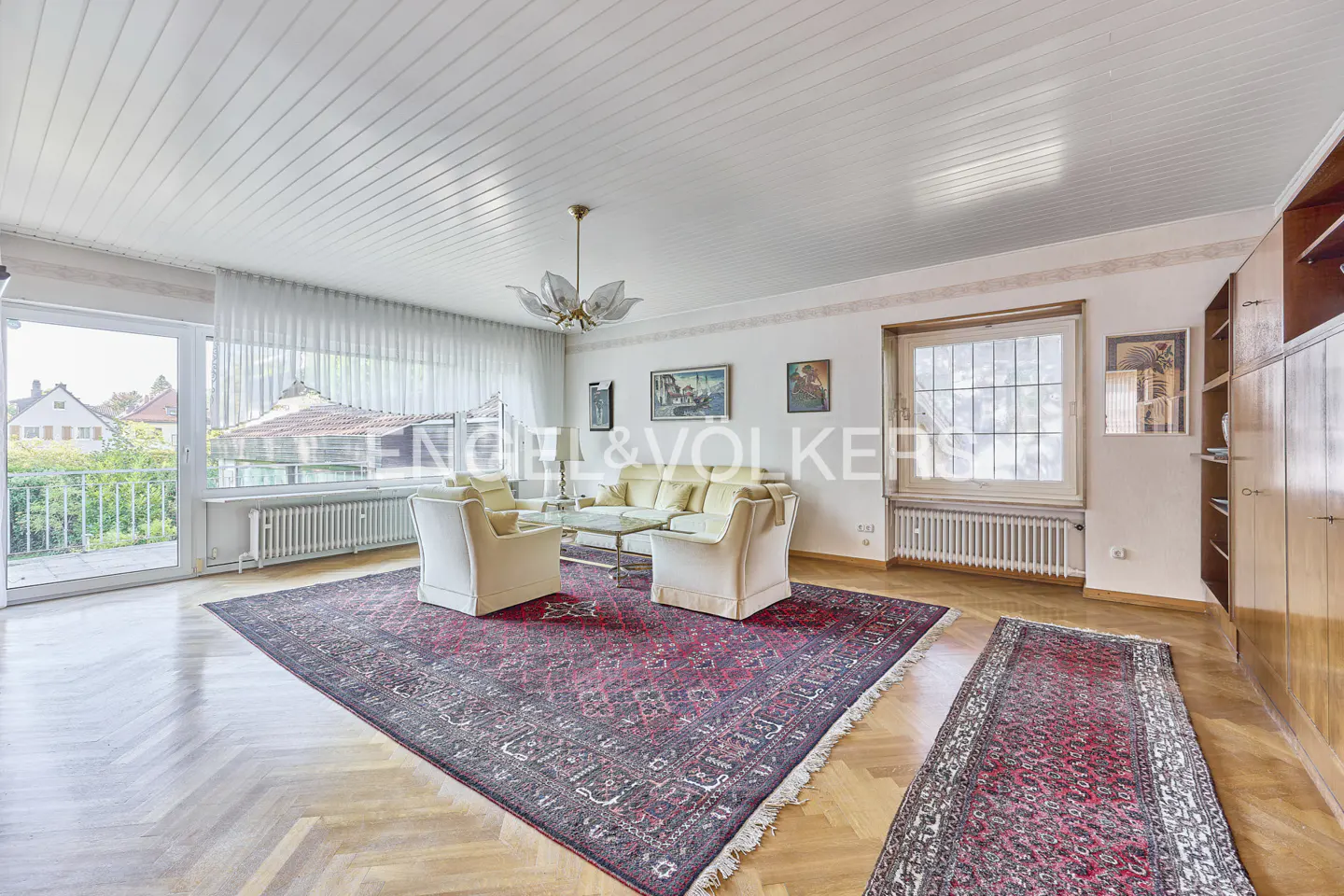Bright living room with herringbone wood floors, cream sofas, a large red patterned rug, and a glass-topped coffee table.