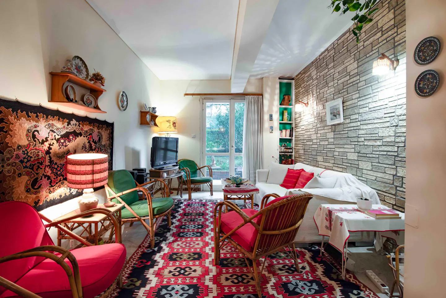 Cozy living room with red and green rattan chairs, a white sofa with red pillows, and a patterned rug. A stone wall and shelves add texture.