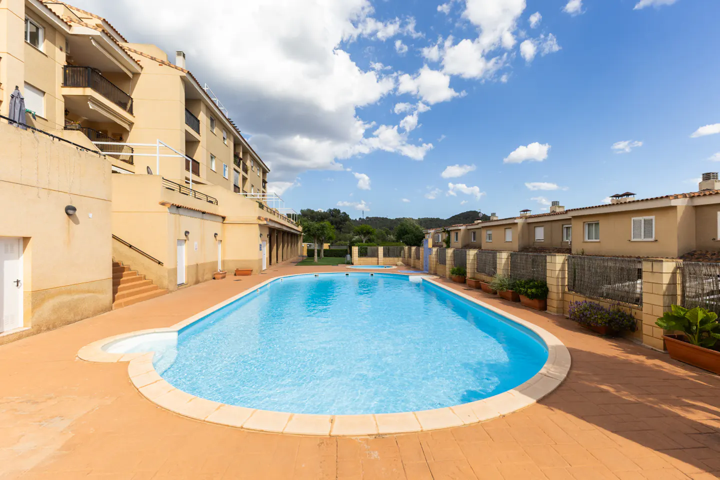 Outdoor pool with clear blue water, surrounded by tan brick patio and buildings under a blue sky with white clouds.