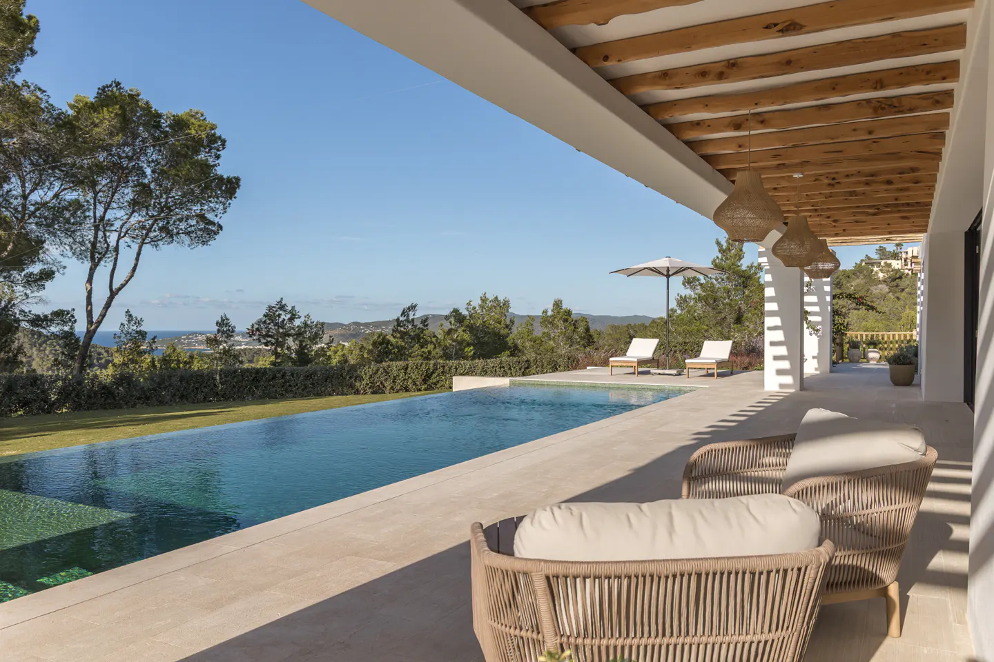 Outdoor patio with wicker chairs, a pool, and lounge chairs overlooking a green landscape and blue sky.