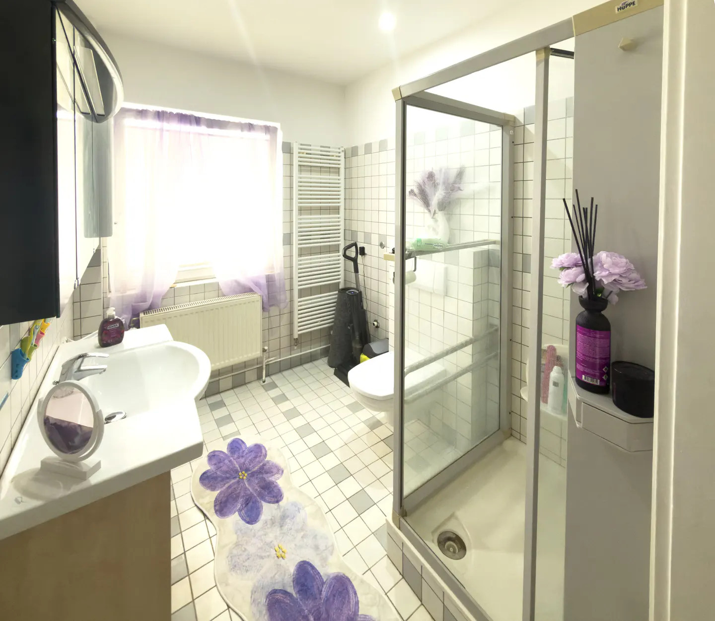 Bright bathroom with white tiled walls, a glass shower, and a white sink. A purple flower rug sits on the gray tiled floor.
