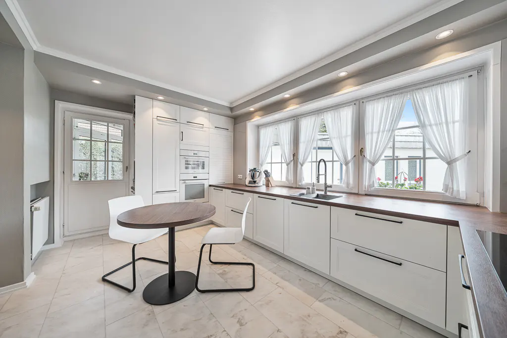 Bright kitchen with white cabinets, wood countertops, and marble floors. A round table with white chairs sits near a door. Large windows with sheer curtains.