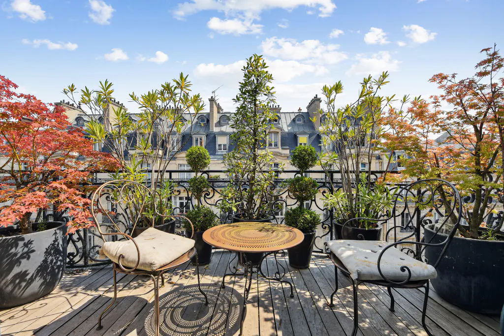 Parisian balcony with wrought iron furniture, potted plants, and city views. Two chairs flank a round table on a wooden deck.