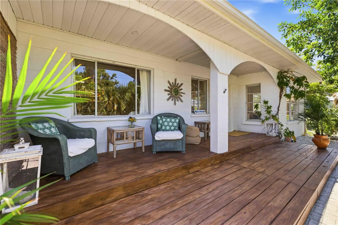 Inviting front porch with wood flooring, wicker chairs with cushions, and small tables. White exterior with arched entryway and lush greenery.