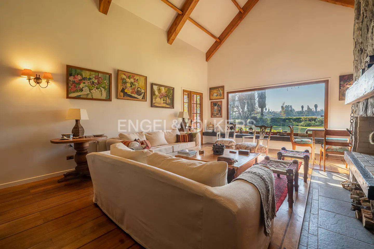 Bright living room with wood floors, white sofas, and a stone fireplace. Large window overlooks a green landscape. Paintings adorn the walls.
