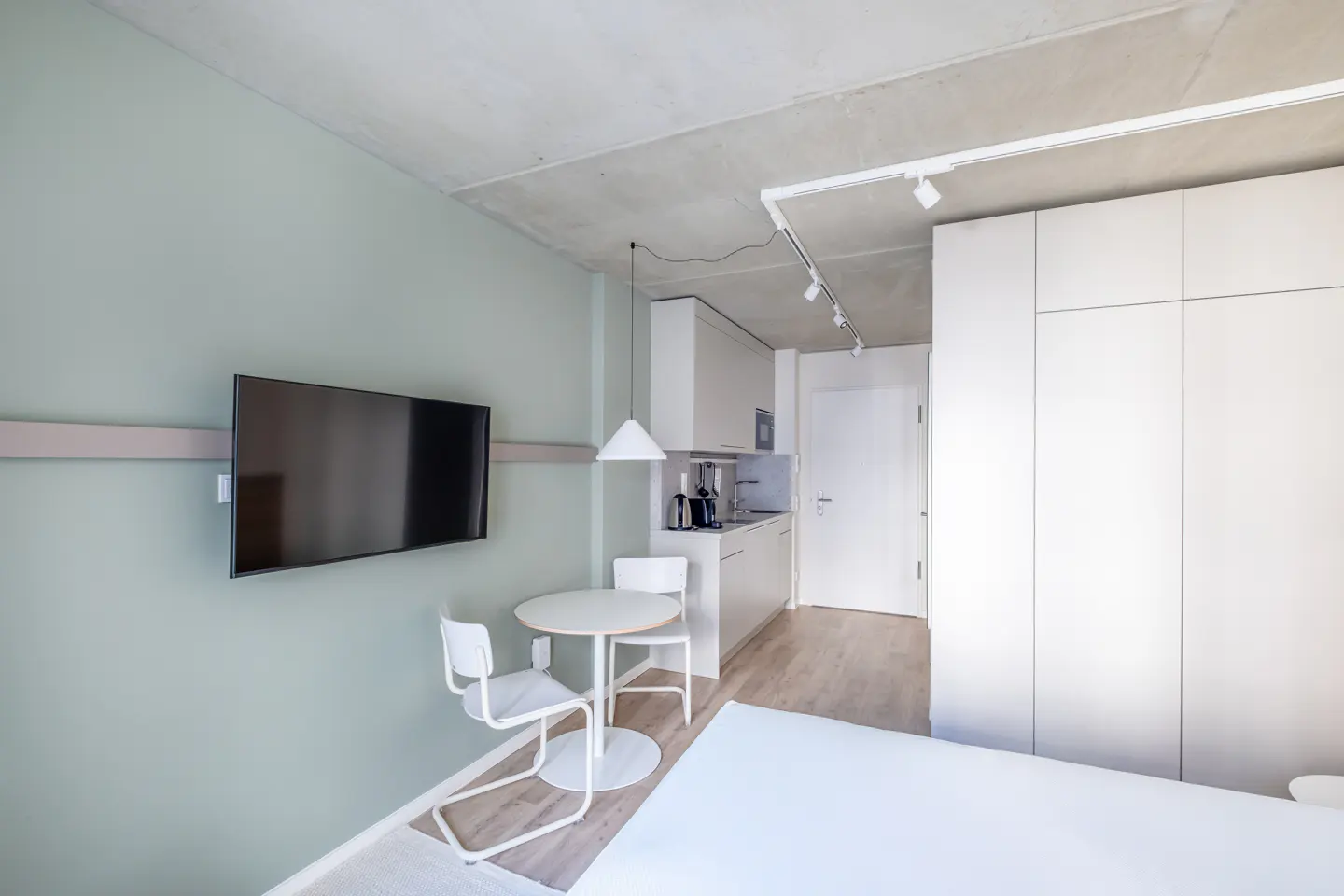 Studio apartment with a TV on a light green wall, a white table and chairs, and a kitchenette with white cabinets.