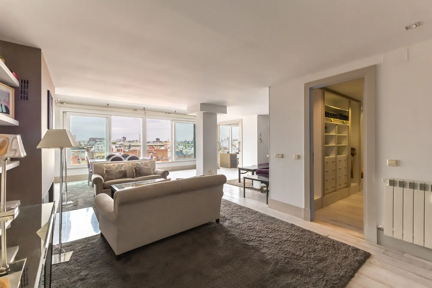 Bright, modern living room with beige sofas, a gray rug, and large windows overlooking a city. A walk-in closet is visible.