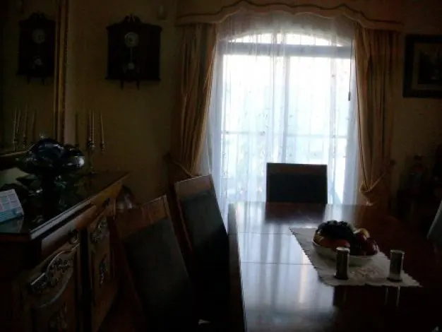 A dimly lit dining room with a long wooden table, chairs, and a fruit bowl. Curtains frame a bright window.