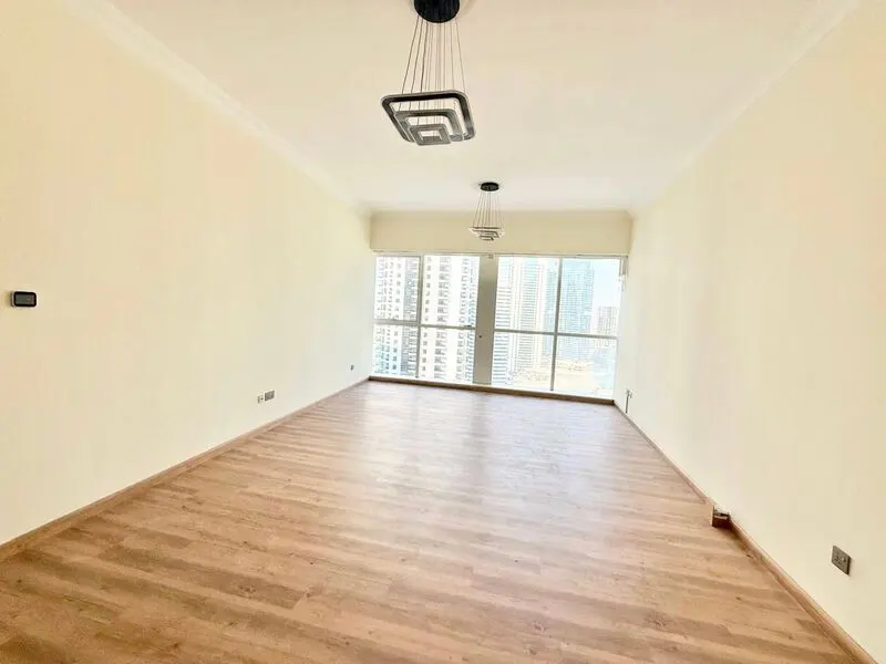 Empty, bright room with light wood floors, cream walls, two modern pendant lights, and floor-to-ceiling city view windows.