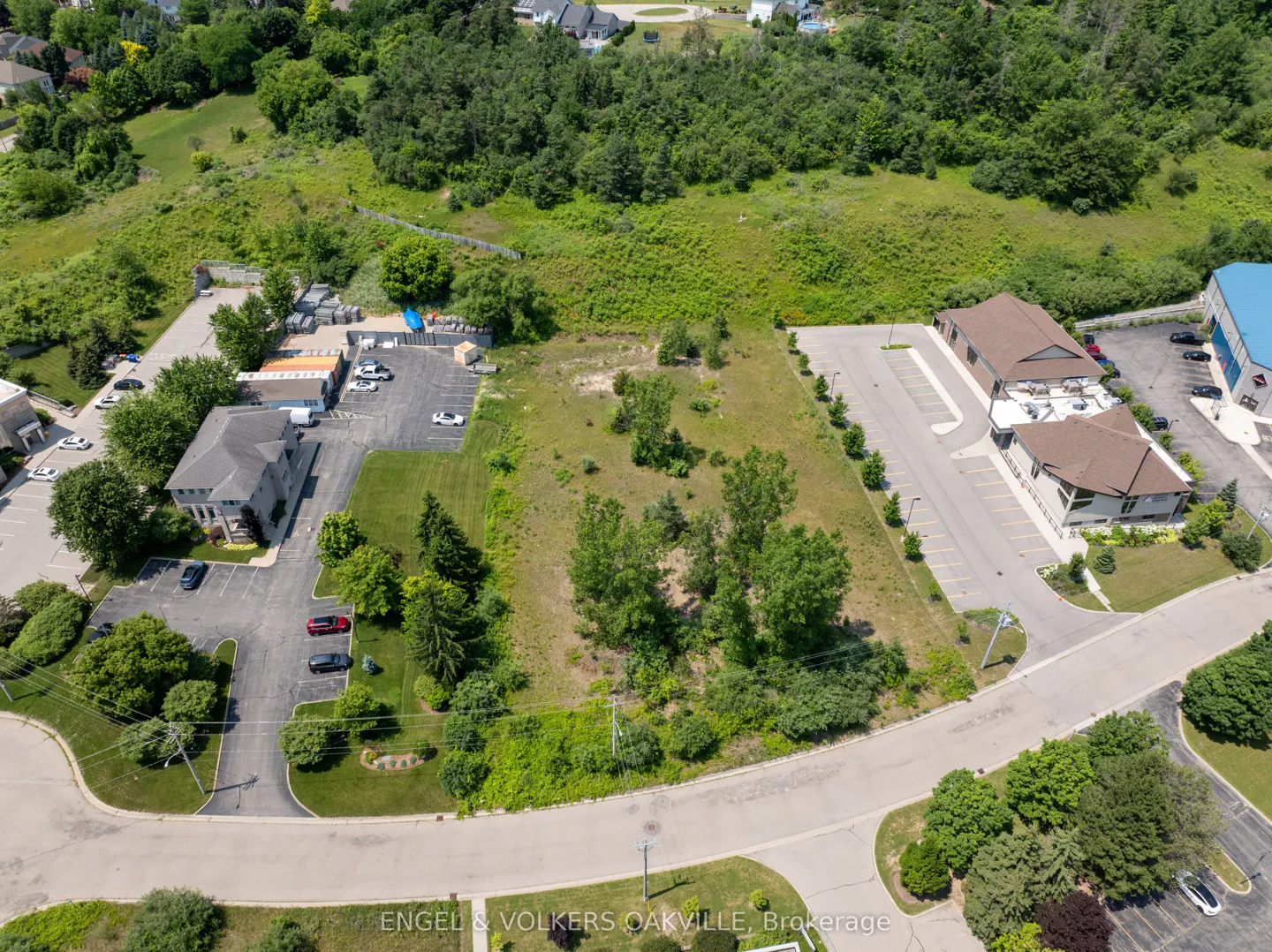 Aerial view of a vacant lot with trees, adjacent to commercial buildings and parking lots, surrounded by green foliage.