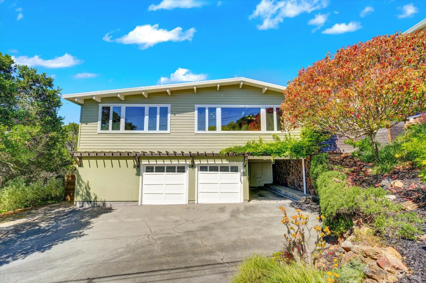 Two-story house with light green siding, white trim, and a two-car garage. A tree with red leaves is on the right. Blue sky.