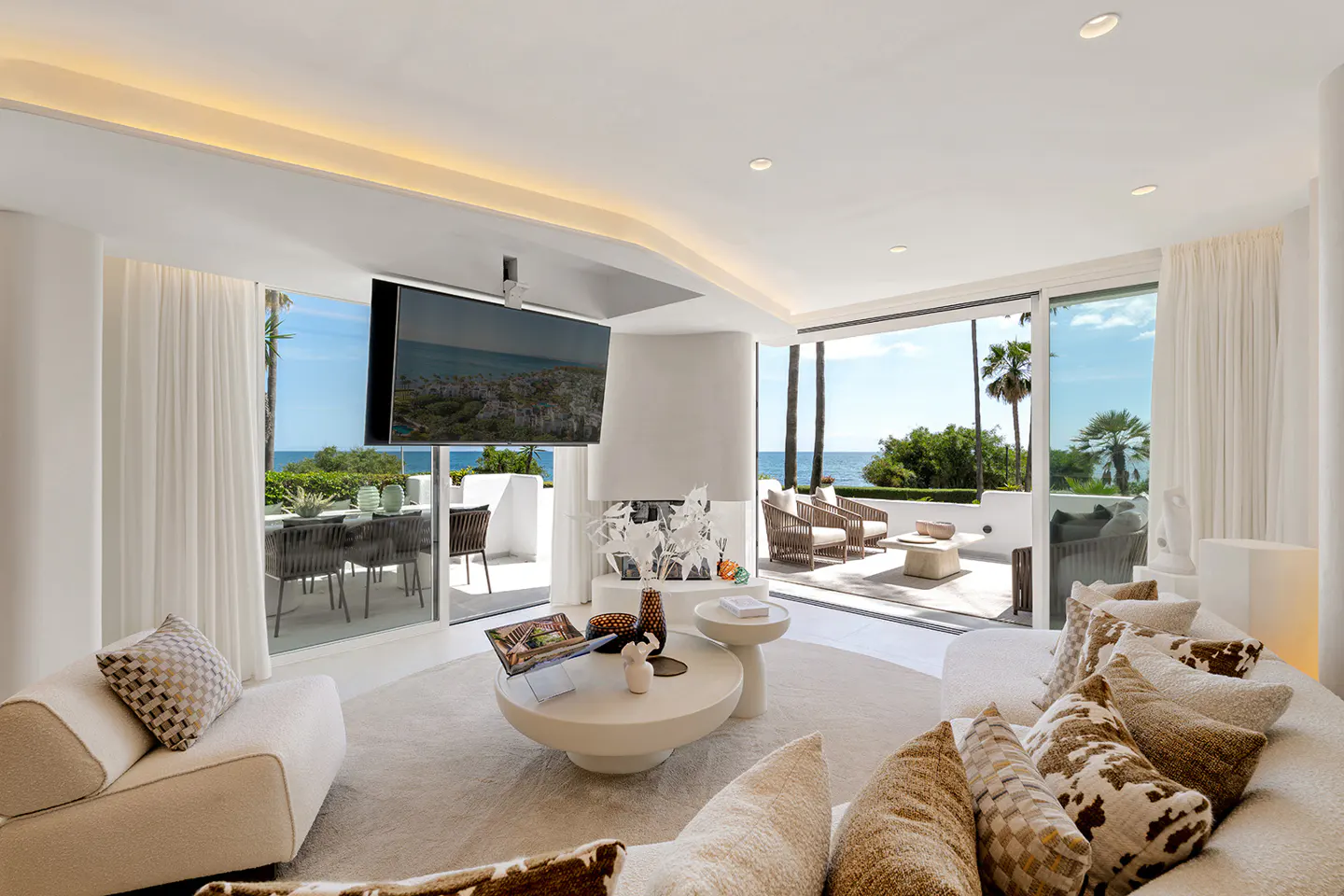 Modern white living room with a curved sofa, round coffee table, and large glass doors opening to a sunny ocean view patio.