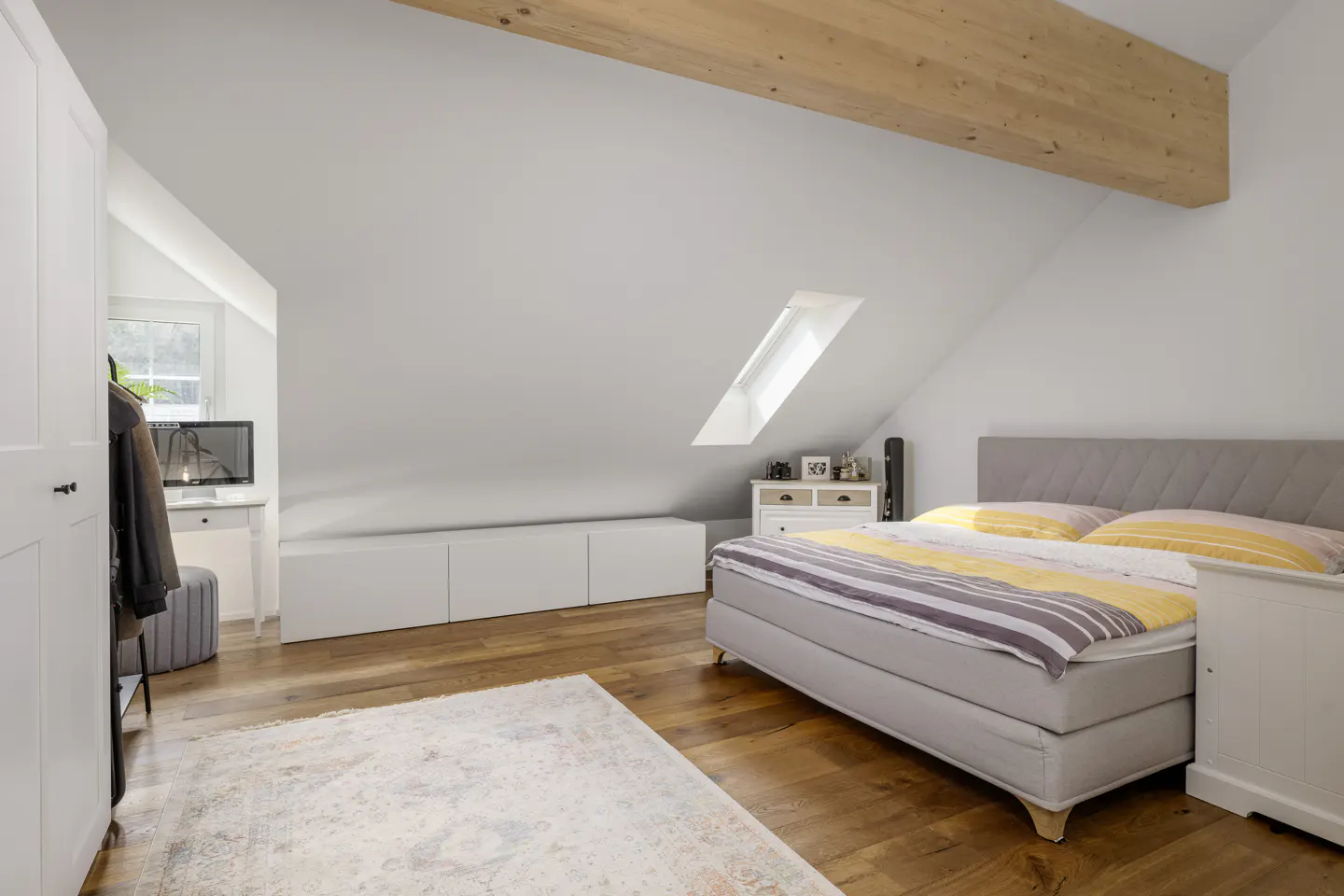 Attic bedroom with wood floors, white walls, and a wooden beam. A bed with yellow and gray bedding sits near a white nightstand and dresser. A skylight is visible.
