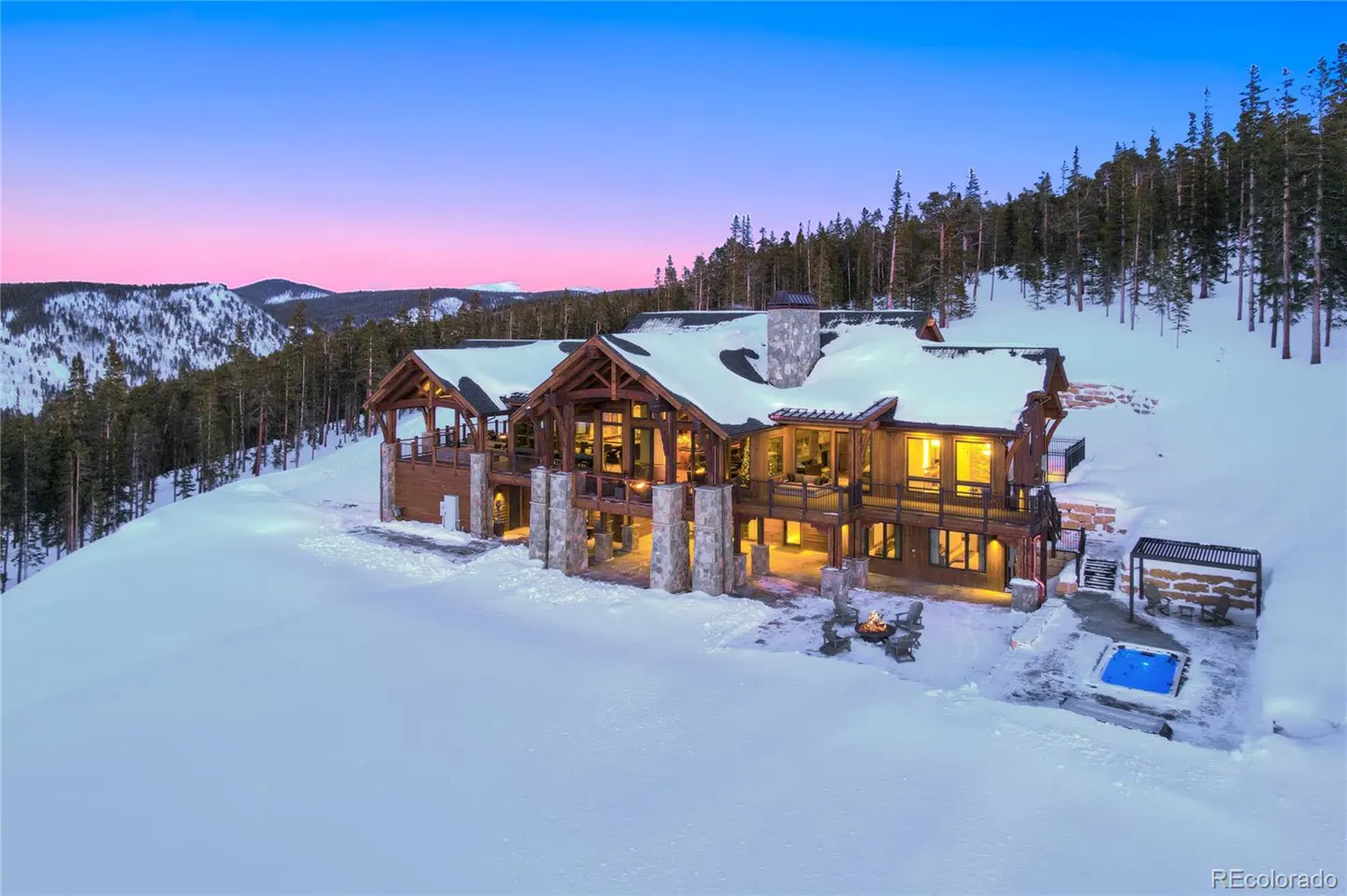 A large, wood and stone mountain home sits on a snowy hillside at dusk, with a hot tub and fire pit.