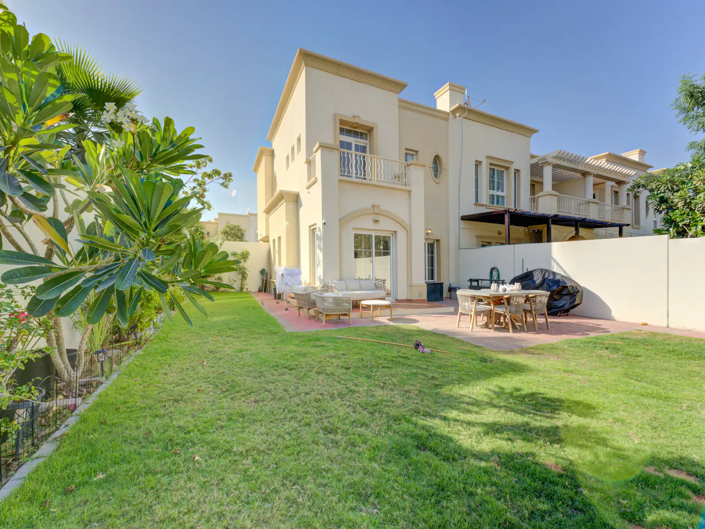 Two-story beige house with a green lawn, patio furniture, and a dining table. The sky is blue and clear.