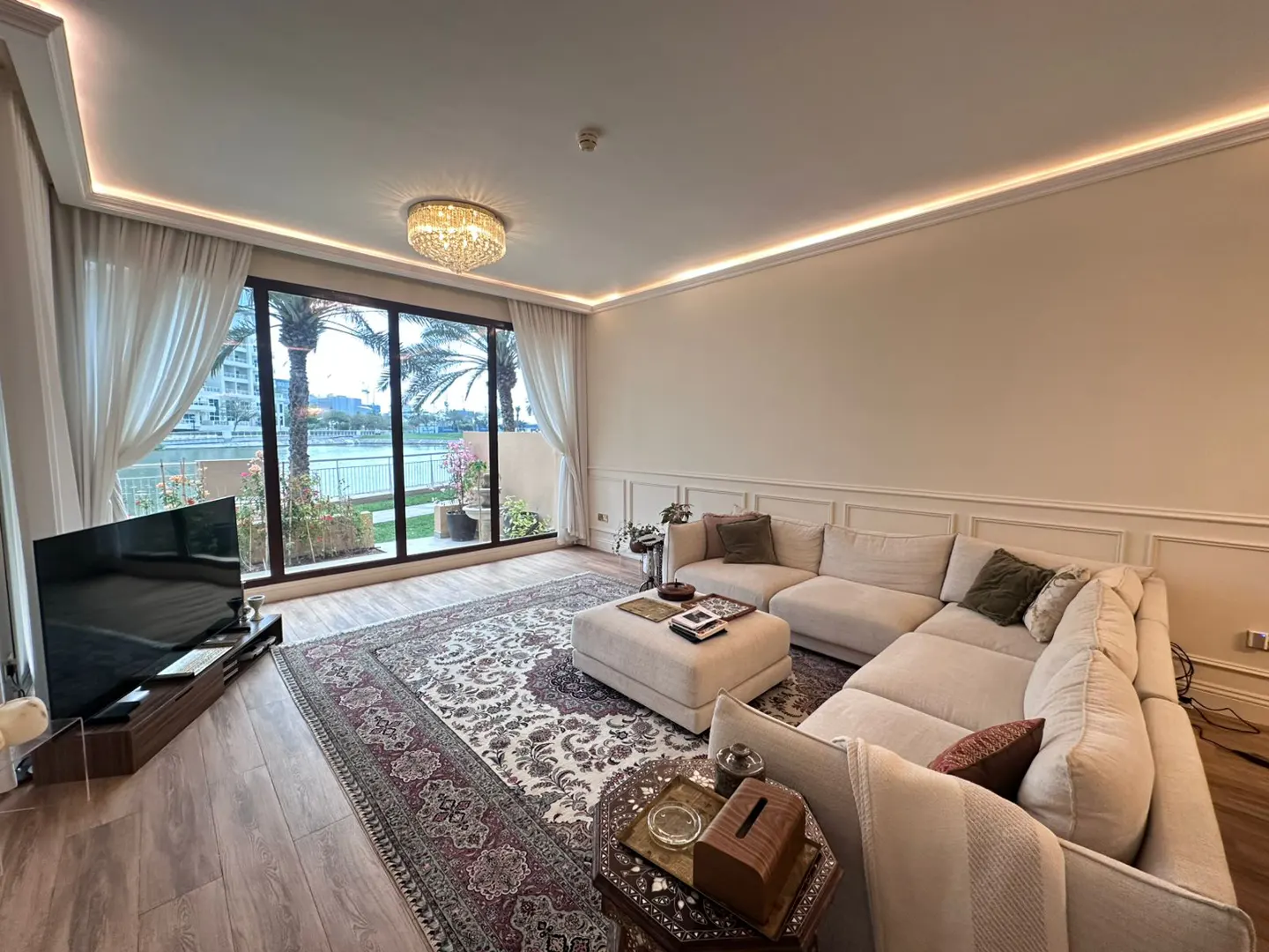 Living room with a large window view of palm trees and water. Beige sectional sofa, patterned rug, and crystal chandelier.