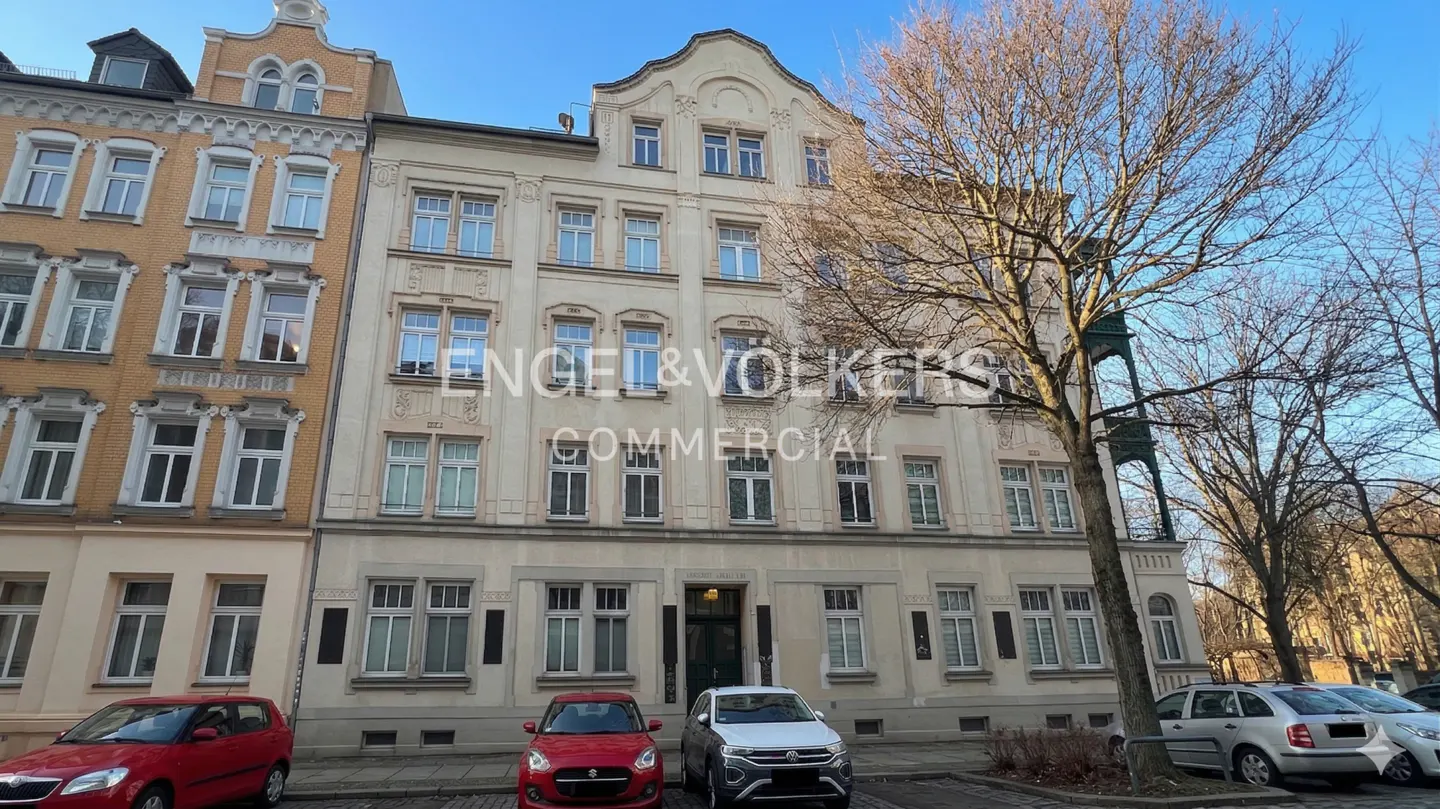 Exterior of a light beige commercial building with many windows and parked cars in front. Engel & Volkers logo visible.
