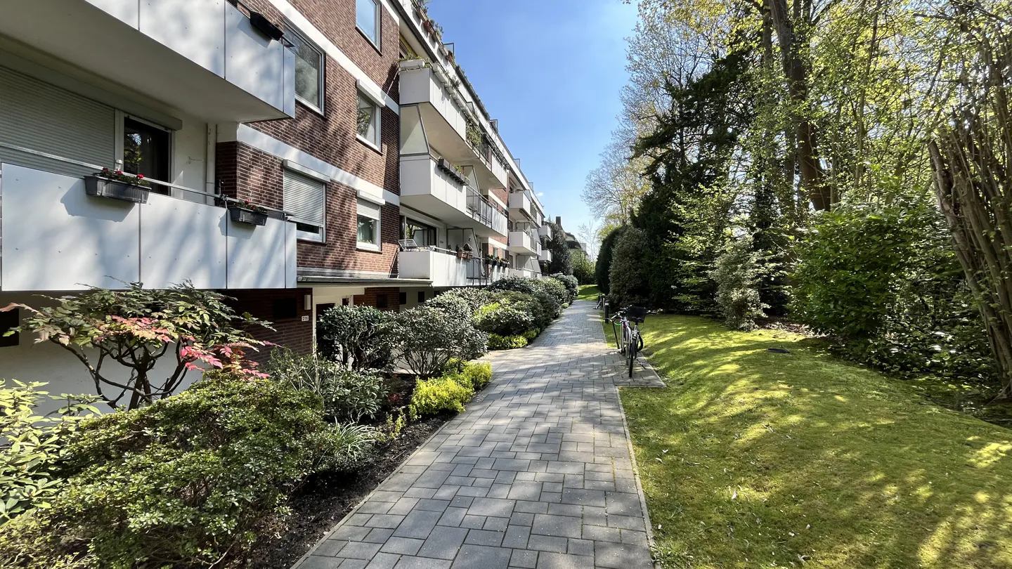 Brick apartment building with white balconies and a gray brick walkway lined with green bushes and trees.