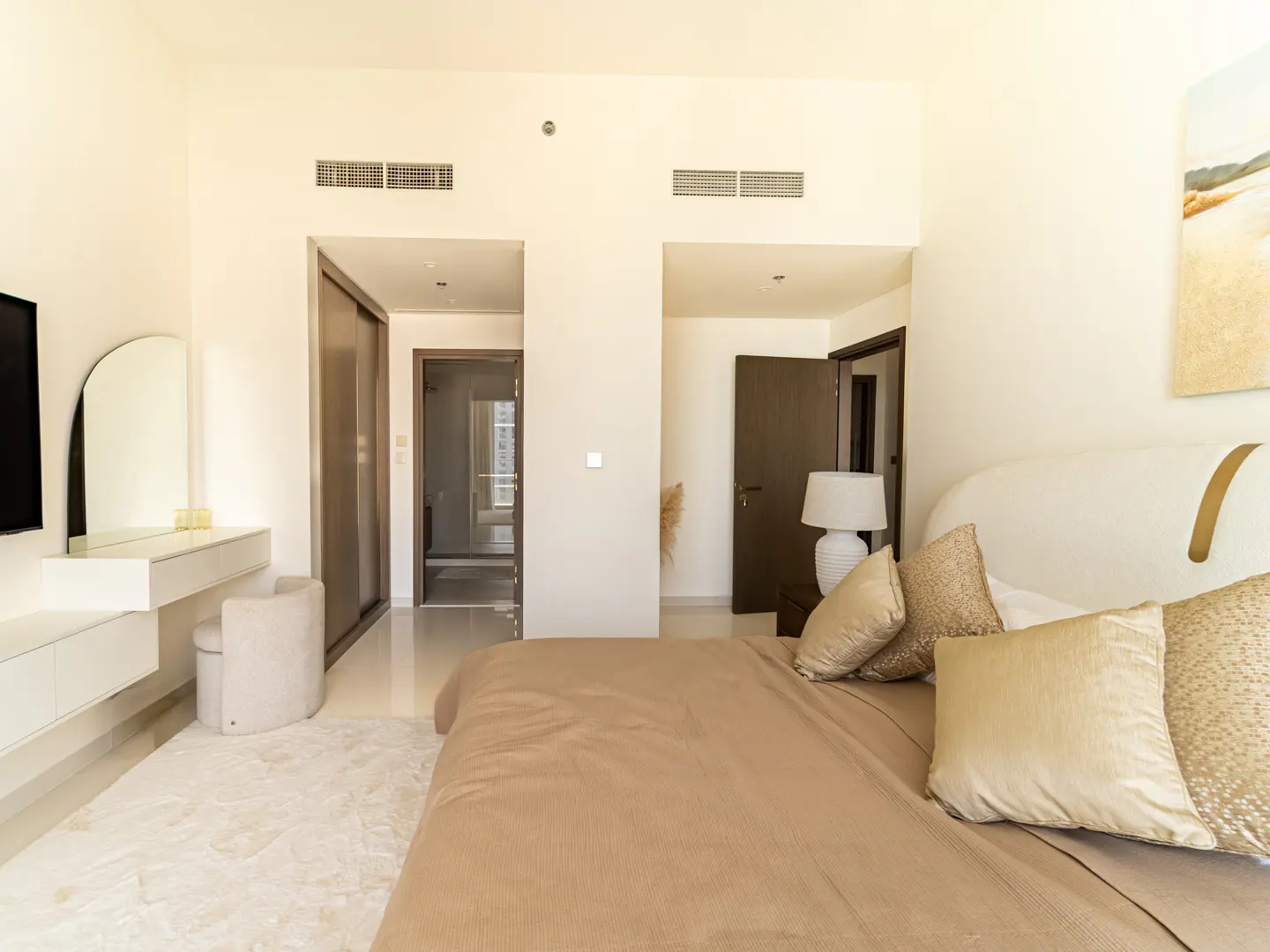 Modern bedroom with a beige bed, gold pillows, white vanity with mirror, and doorways leading to a closet and bathroom.